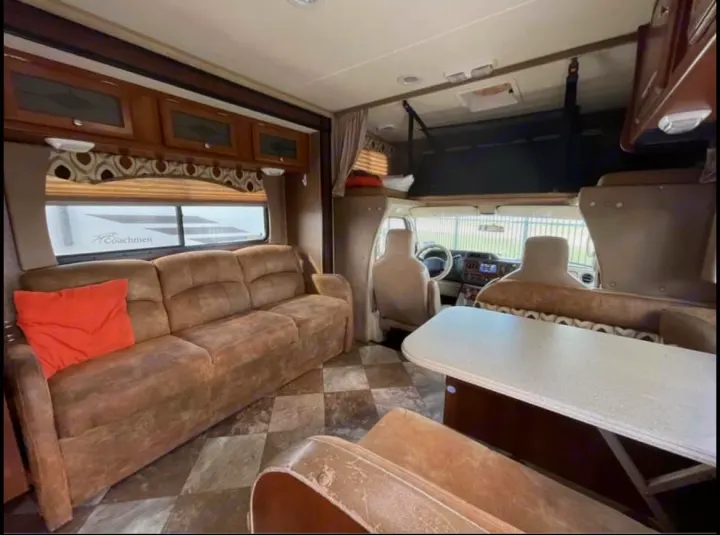 View of an RV rental's living space, featuring a tan sofa, dining table, and front cabin with an overhead bunk.. Coachmen Leprechaun 2015