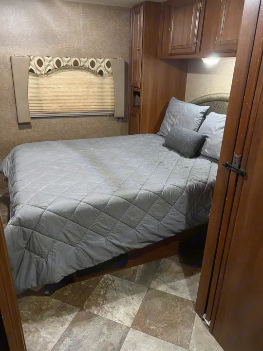 Cozy RV rental bedroom with gray bedding, built-in cabinets, and patterned window treatments, visible from an open doorway.. Coachmen Leprechaun 2015