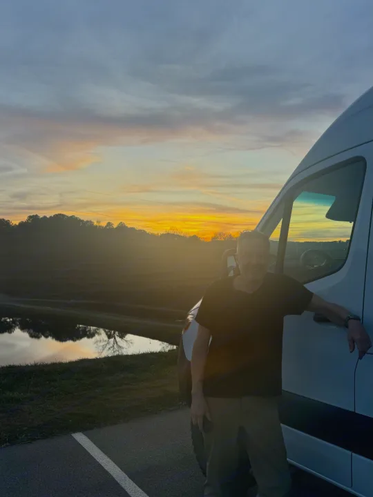 Man smiles next to an RV rental van at sunset, overlooking a serene lake. Ideal for peaceful getaways.. Thor Tranquility 2023