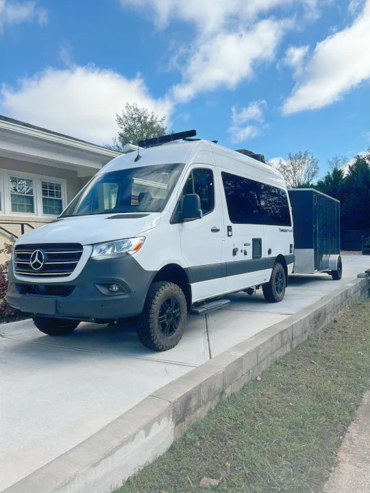 Bright white Mercedes Sprinter camper van, likely an rv rental, with a black trailer parked on a driveway under a blue sky.. Thor Tranquility 2023
