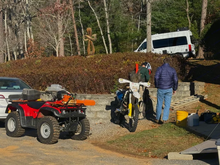 White Tranquility 4x4 RV rental, red ATV, and dirt bike parked in a wooded area as two men prepare for off-road riding.. Thor Tranquility 2023