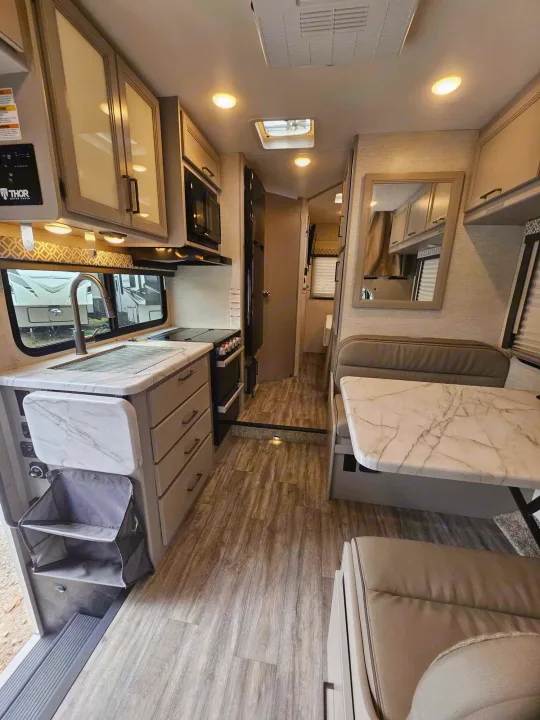 Explore this spacious RV rental interior, featuring a modern kitchen, comfortable dining area, and light wood-look flooring.. Thor Motor Coach Other 2024