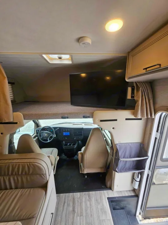 Spacious RV rental interior with bright ceiling light, over-cab bed, cockpit, TV, and living area.. Thor Motor Coach Other 2024