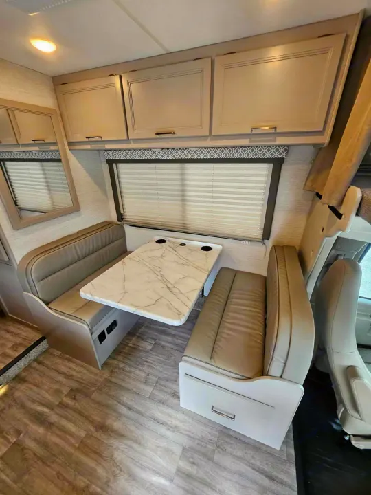 Cozy RV rental dinette with light upholstered bench seating, a stylish marble-patterned table, and ample overhead cabinets.. Thor Motor Coach Other 2024