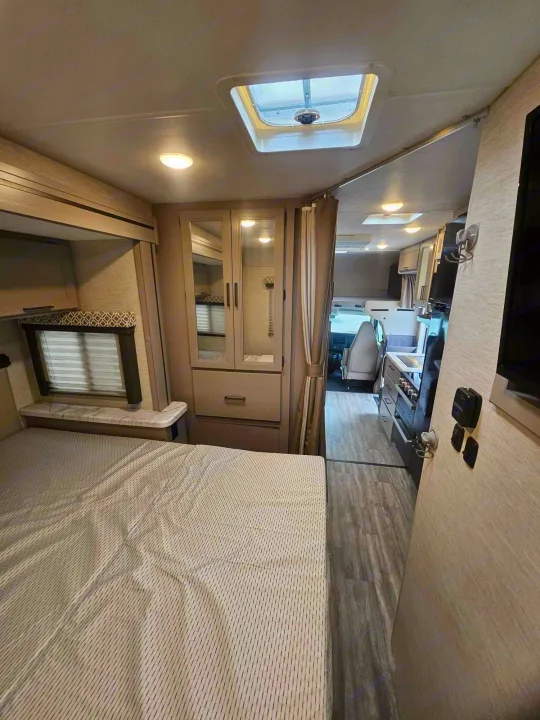 Inside an RV rental: a comfortable bed, window, and mirrored wardrobe, with a skylight above. View extends to kitchen and cockpit.. Thor Motor Coach Other 2024