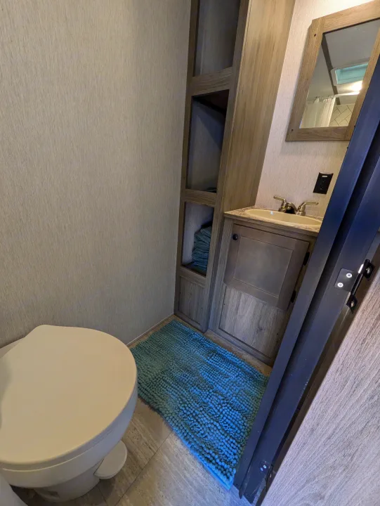Functional RV rental bathroom: white toilet, vanity with sink, mirror, open shelves with towels, & blue bath mat.. Sunset Park & Rv Inc. 19rb 2025