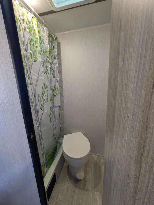 Compact RV rental bathroom with a shower (birch tree curtain) and toilet. Features light grey textured walls and wood-look flooring.. Sunset Park & Rv Inc. 19rb 2025