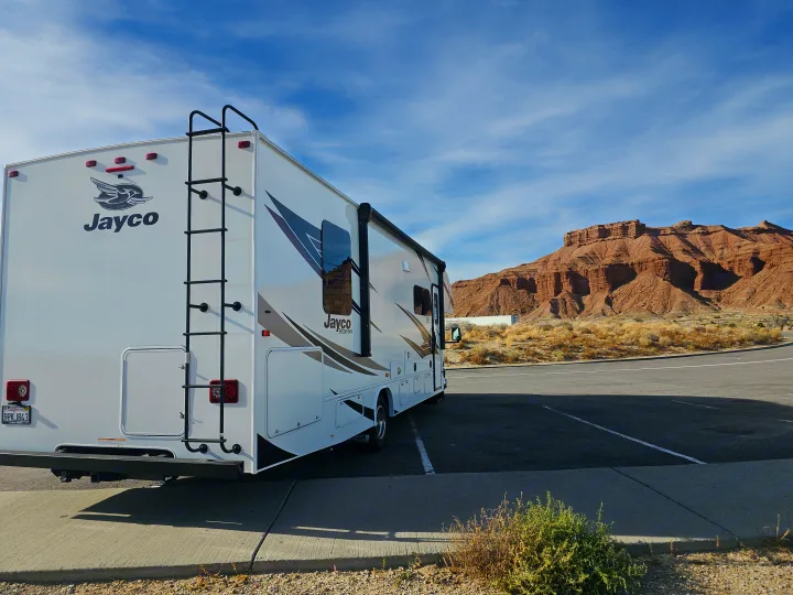 A white Jayco RV rental is parked, with striking red rock mountains under a blue sky in the background.. Jayco Redhawk 2024