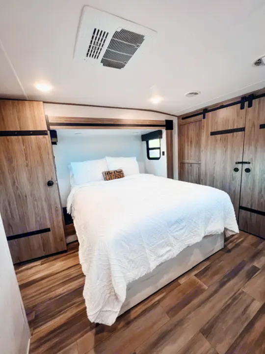 This RV rental bedroom features a cozy bed with white bedding, wood-look floors, and built-in storage.. Heartland RVs Milestone 2021