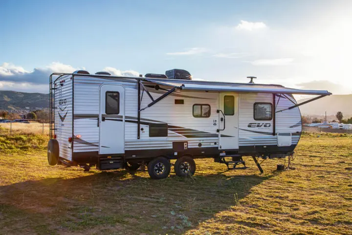 White travel trailer with extended awning, perfect for an RV rental, parked in a sunny grassy field with distant hills.. Forest River Evo 2019