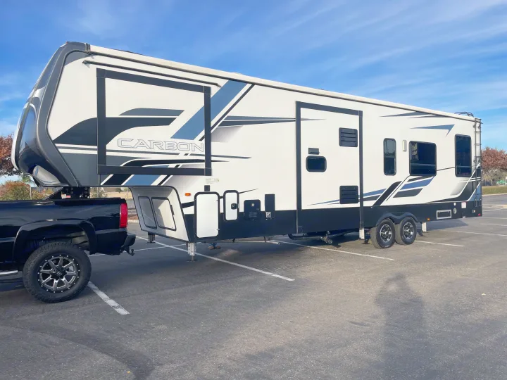 Black pickup truck towing a large white and black Carbon fifth-wheel RV rental in a parking lot.. Keystone Carbon 2021