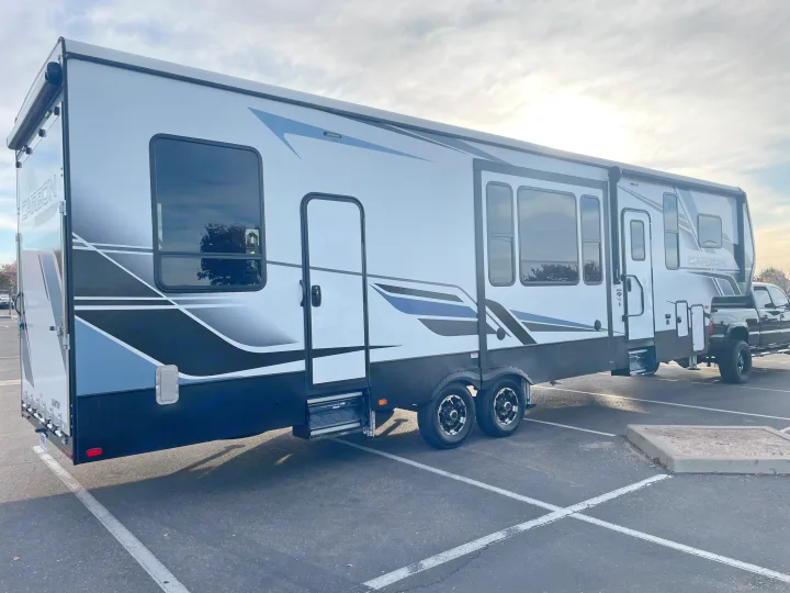 Large white Carbon toy hauler fifth wheel RV, possibly an RV rental, attached to a black pickup truck in a parking lot.. Keystone Carbon 2021