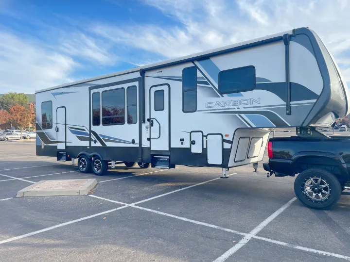 A large white Carbon fifth-wheel RV rental, hitched to a black truck, is parked in an asphalt lot on a sunny day.. Keystone Carbon 2021