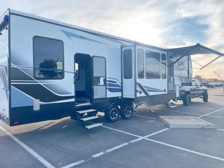 Large white and black fifth-wheel RV rental, with slides out and awning deployed, hitched to a truck in a parking lot. Ready for adventure.. Keystone Carbon 2021