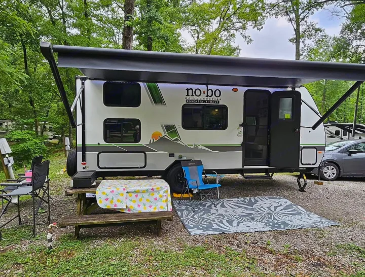 Enjoying an RV rental experience: a modern camper with awning, picnic table, and outdoor seating at a forested campsite.. No Boundaries NoBo 19.3 Essentials Only 2024