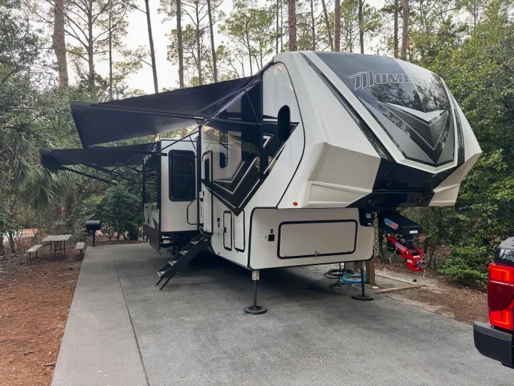 Modern black and white fifth-wheel RV with extended awnings parked at a scenic wooded campsite, ready for your rv rental trip.. Grand Design Momentum M-Class Toy Hauler 2023