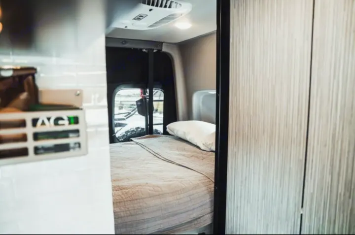 Cozy rv rental interior: a comfortable bed with a quilted blanket and pillow, and an AC unit in a compact camper van.. Winnebago Revel 2022