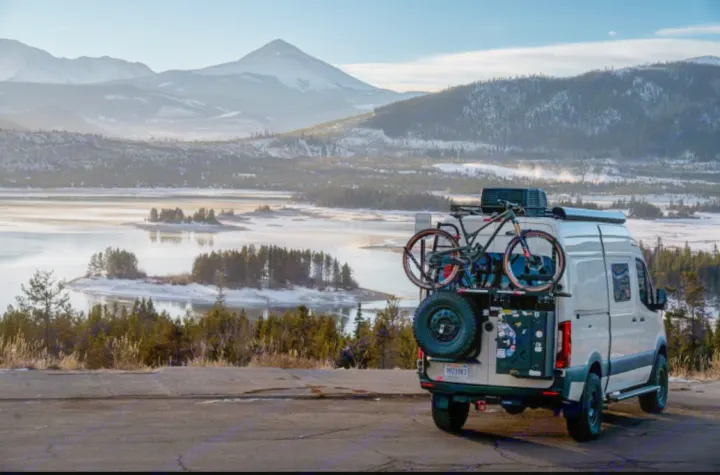 An RV rental camper van, equipped with bikes, overlooks a picturesque frozen lake and majestic snow-capped mountains.. Winnebago Revel 2022