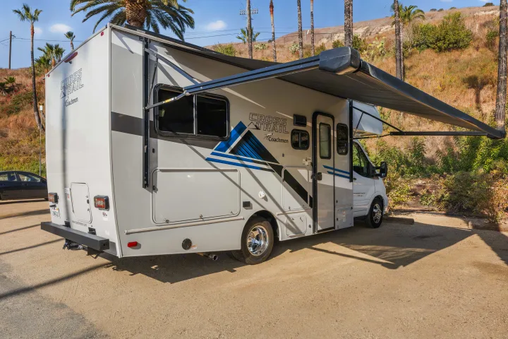 Light gray Coachmen Cross Trail RV rental with an extended awning, parked on a dirt lot by palm trees near the ocean.. COACHMEN Ford Transit 2022