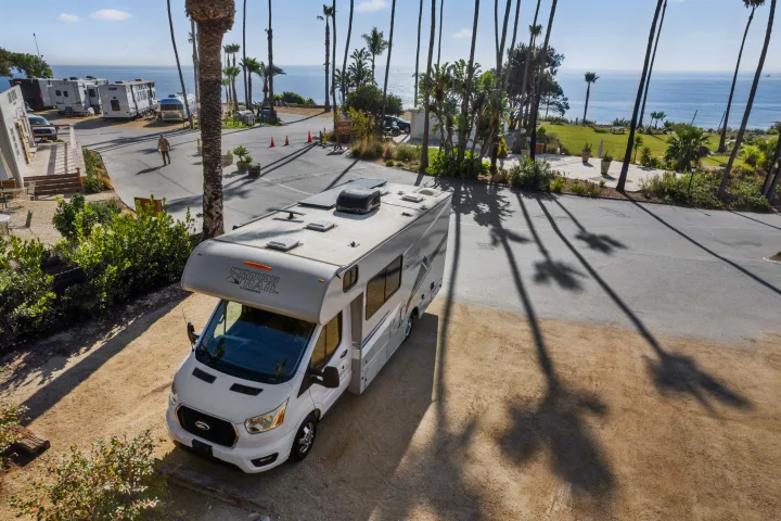 A white Cross Trail RV rental is parked at a sunny coastal RV campground with palm trees & ocean views.. COACHMEN Ford Transit 2022
