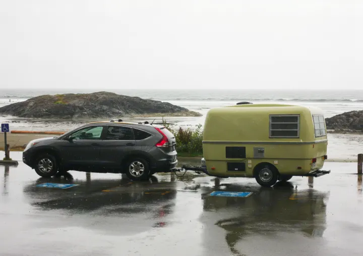 Grey SUV with an rv rental on a rainy coastal road trip. The light green camper is parked near a rocky coast and ocean.. Trillium Rv 1300 1976