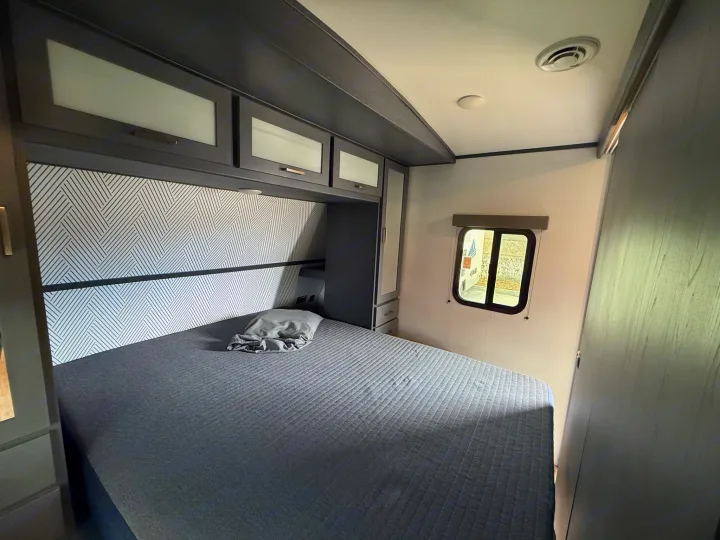 Inside an **RV rental**, a cozy bedroom features a gray quilted bed, patterned headboard, and ample overhead storage.. Keystone Bullet 2022