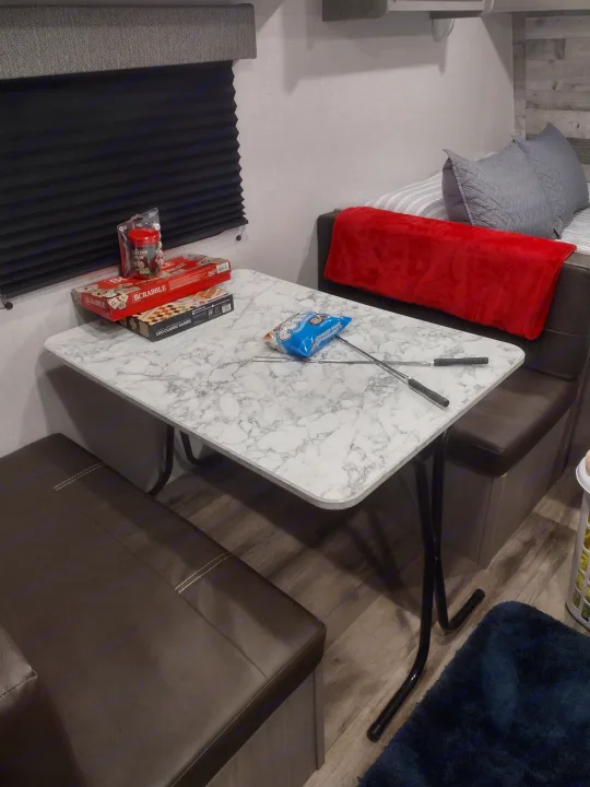 RV rental dining area: marble table with board games, marshmallows, and roasting sticks. Bed with red blanket in background.. Coleman Lantern 2023
