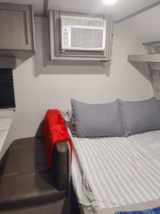 An RV rental interior showing a compact sleeping area with a mounted AC unit, bed with pillows, and a red blanket on a bench.. Coleman Lantern 2023
