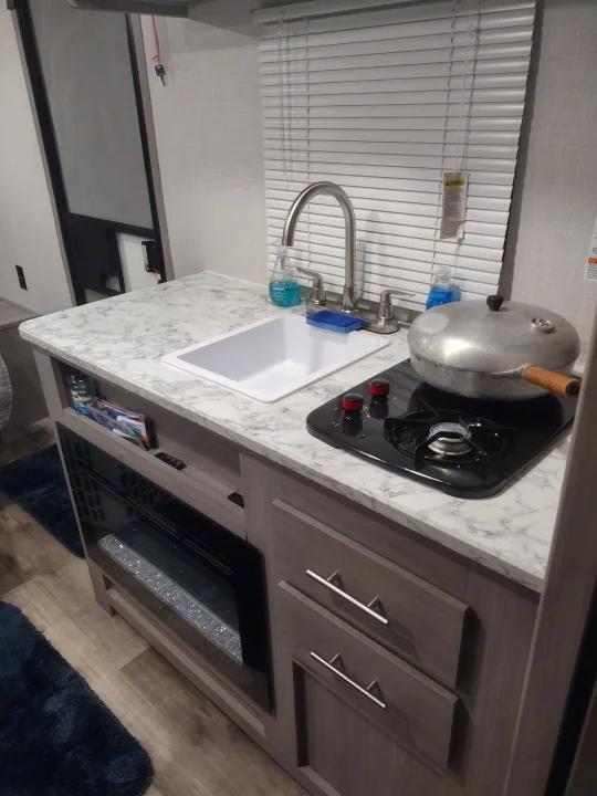 View of a modern RV rental kitchen interior, showing a countertop with sink, stovetop, and an electric fireplace.. Coleman Lantern 2023