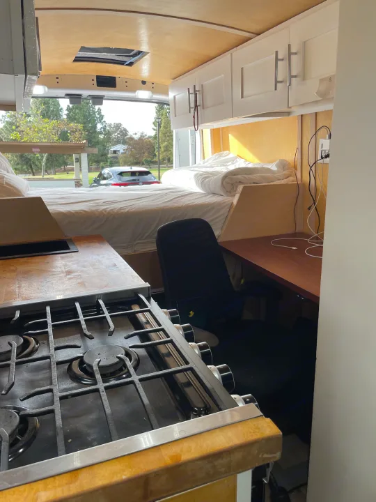 Spacious RV rental interior with a cozy bed, functional kitchen, and dedicated workspace, perfect for mobile living.. Ford Transit 2023