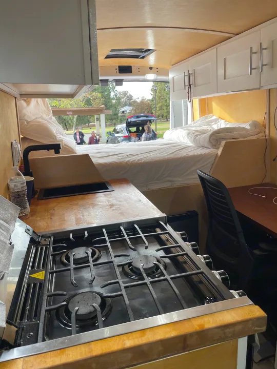 This RV rental showcases a camper van interior with a well-equipped kitchen, bed, workspace, and a view of a park.. Ford Transit 2023