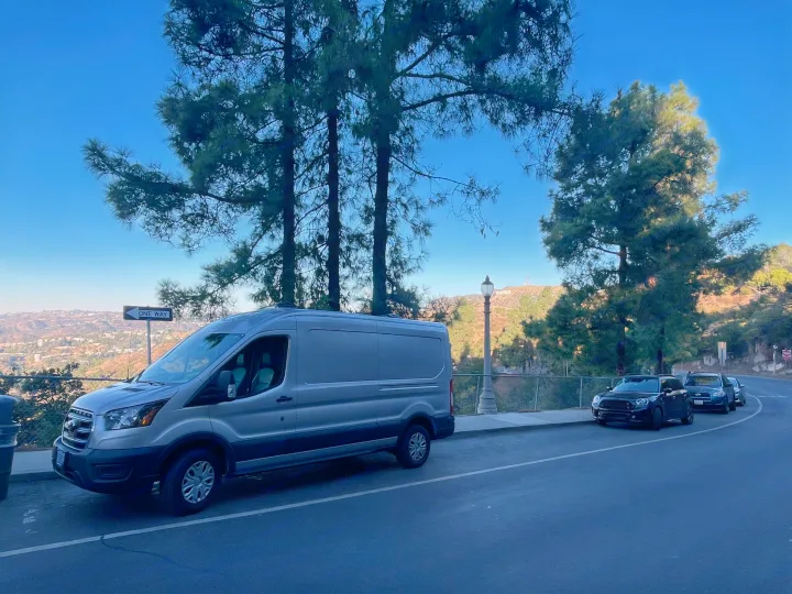 A grey Ford Transit van, suitable for an RV rental adventure, overlooks the Hollywood sign and Los Angeles cityscape.. Ford Transit 2023
