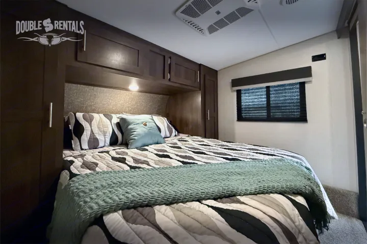 Cozy RV rental bedroom with a bed featuring patterned comforter & green throw, overhead cabinets, window with blinds, and ceiling AC.. KZ Sportster 2021