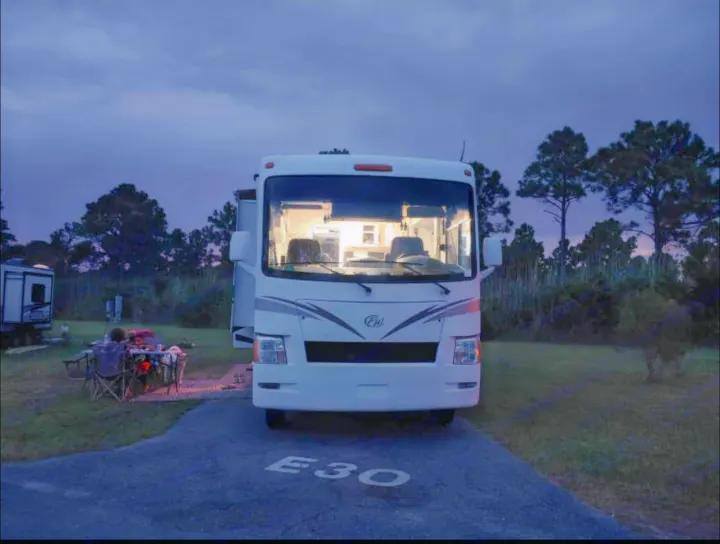 An RV rental motorhome glows at dusk in campsite E30, while people relax by an outdoor table. Serene camping scene.. Thor Motor Coach windsport 31D 2012