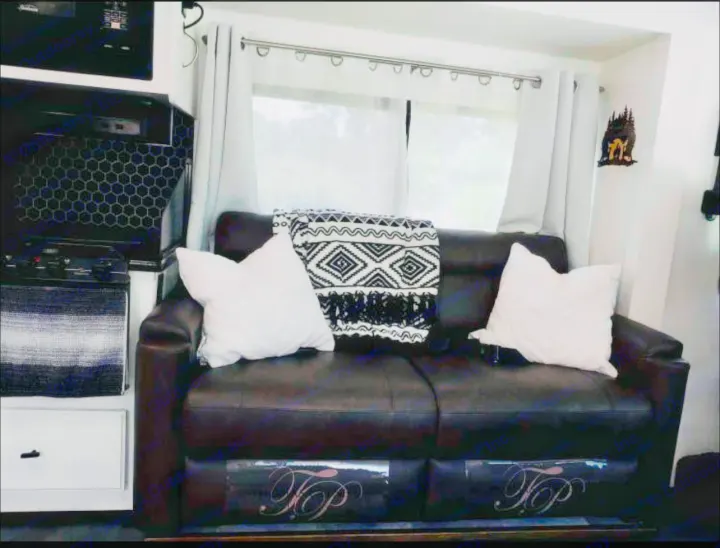 Cozy RV rental living area featuring a dark brown sofa with white pillows, a patterned blanket, and bright curtains by a window.. Thor Motor Coach windsport 31D 2012