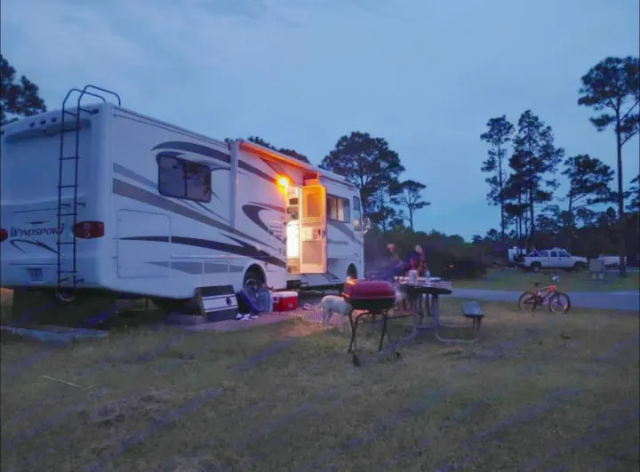 An rv rental lights up a cozy campsite at dusk. People and a dog gather around a grill and picnic table, enjoying the outdoor evening.. Thor Motor Coach windsport 31D 2012