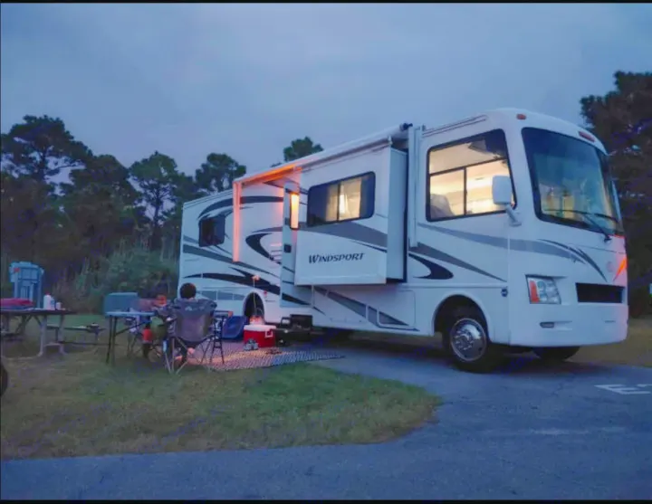 An RV rental parked at a campsite under a twilight sky. Campers relax outside their illuminated motorhome.. Thor Motor Coach windsport 31D 2012