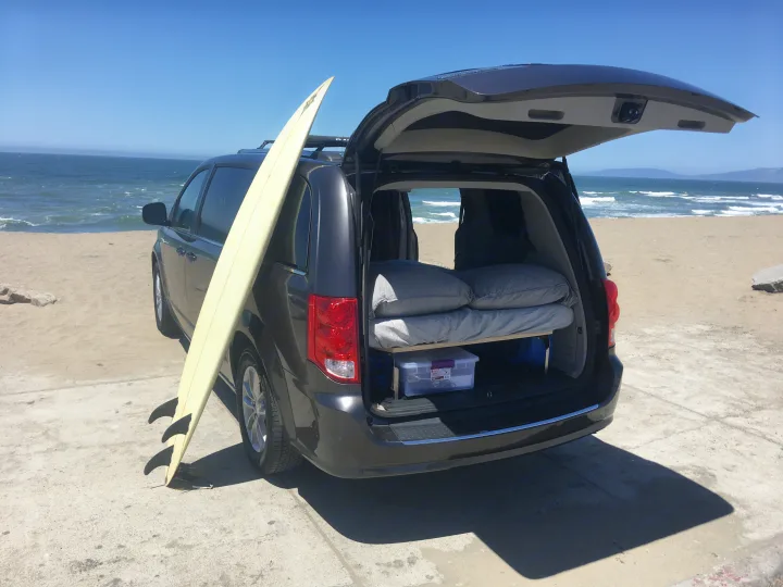 A dark minivan is parked at the beach, its tailgate open revealing a bed and storage. A surfboard leans on the side. Ideal for a compact RV rental trip.. Dodge Caravan 2019