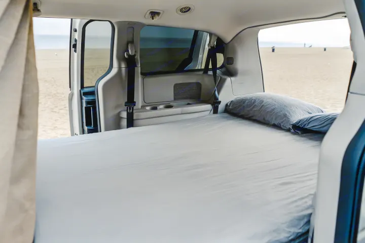 Interior of an **rv rental** minivan, converted with a comfy bed and pillows, parked right on a sandy beach by the ocean.. Dodge Caravan 2019