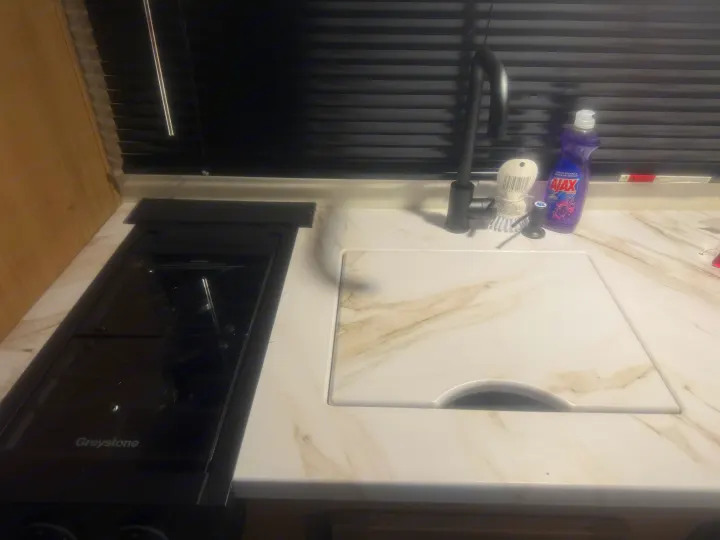 Compact RV rental kitchen features a Greystone stove, black faucet, and sink with cutting board on a marble-look counter.. Forest River Wolf Pup 2025
