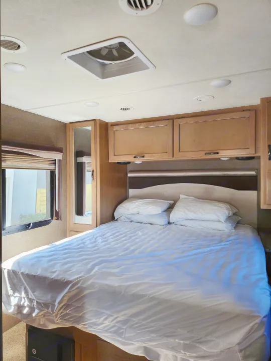 Inside an RV rental, a cozy bedroom with a neatly made bed, white linens, wood cabinets, and a window for natural light.. Thor A.C.E. 2018