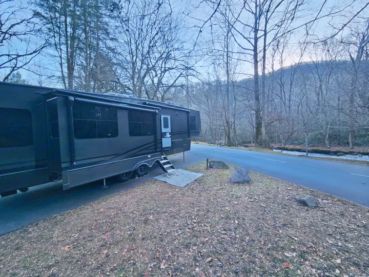 Dark fifth-wheel RV rental parked in a wooded campsite next to a road and stream. Winter trees and mountains.. Keystone RV Cougar 2024