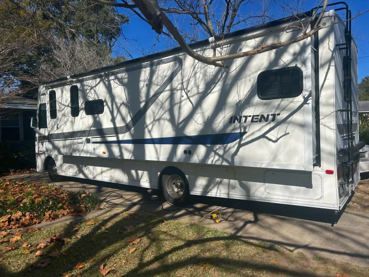 A large white Intent motorhome, possibly an RV rental, is parked outdoors on a sunny day with tree shadows. Electrical cords visible.. Winnebago Intent 2018