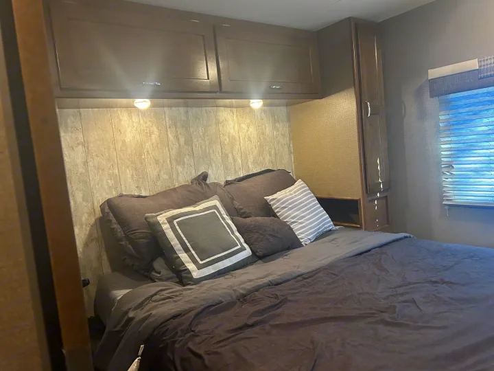Comfortable RV rental bedroom featuring a bed, wood-paneled wall, overhead cabinets, and window. Ideal for trips.. Winnebago Intent 2018