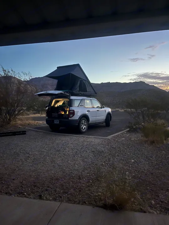 White SUV with rooftop tent deployed, open hatch revealing gear, in a desert campsite at twilight. Consider this RV rental.. Ford Bronco Sport 2022