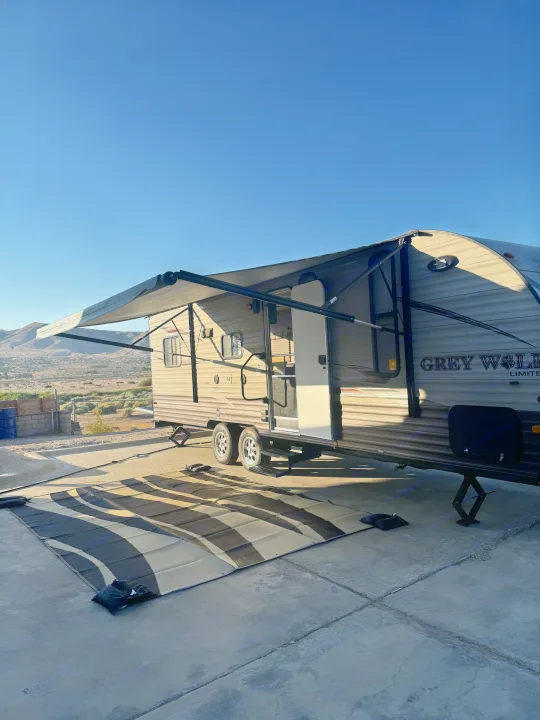 An RV rental with its awning extended and a striped mat laid out on concrete, ready for a sunny outdoor retreat.. Forest River Cherokee Grey Wolf 2016