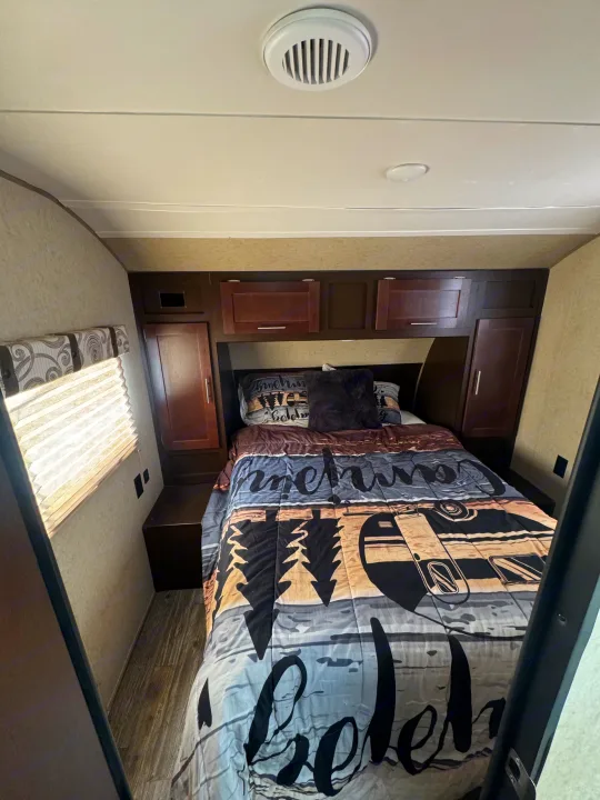 Compact RV rental bedroom featuring a bed with nature-themed bedding, dark wood cabinets, and a window.. Forest River Cherokee Grey Wolf 2016
