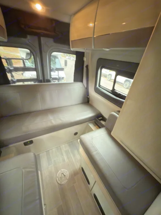 View inside an RV rental: two long grey cushioned benches facing each other, faux wood floor, and side windows.. Thor Tranquility 2023