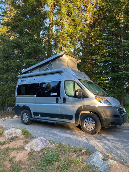 A silver pop-top RV rental camper van is parked on a paved spot among tall evergreen trees, perfect for camping.. Thor Scope 2023