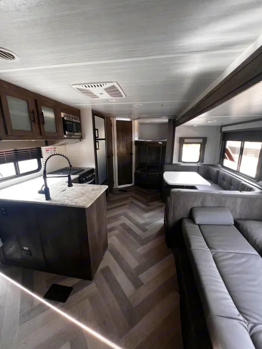Modern rv rental interior featuring kitchen, dining booth, gray sofa, and bunk beds on a herringbone floor.. Forest River Wildwood 2022